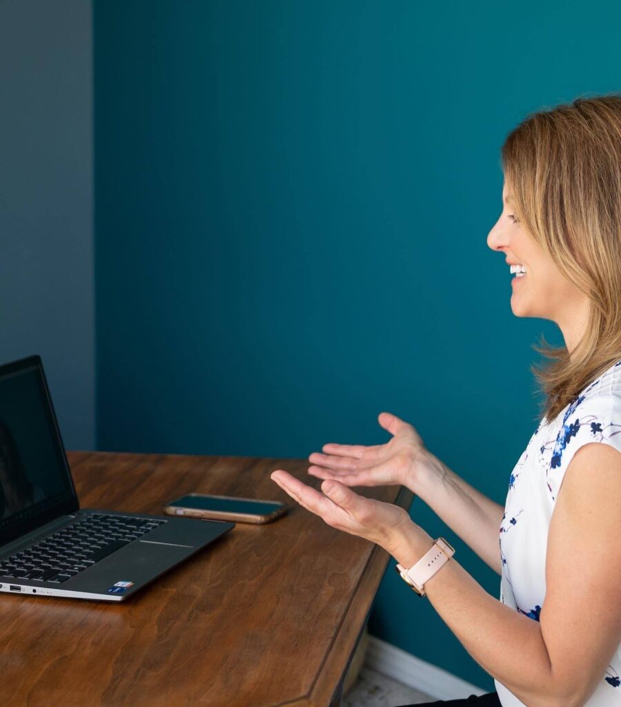 Woman smiling and gesturing while talking to her laptop during a virtual meeting, sitting at a wooden desk against a teal wall.