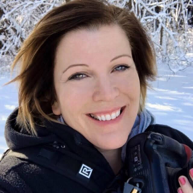 Woman with shoulder-length hair smiling outdoors in winter, holding a camera and wearing a jacket.