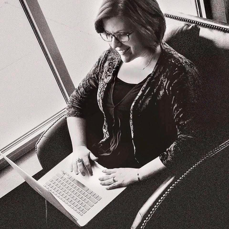 Woman sitting on a chair by a window smiling while typing on a laptop, representing focused work and business reflection.
