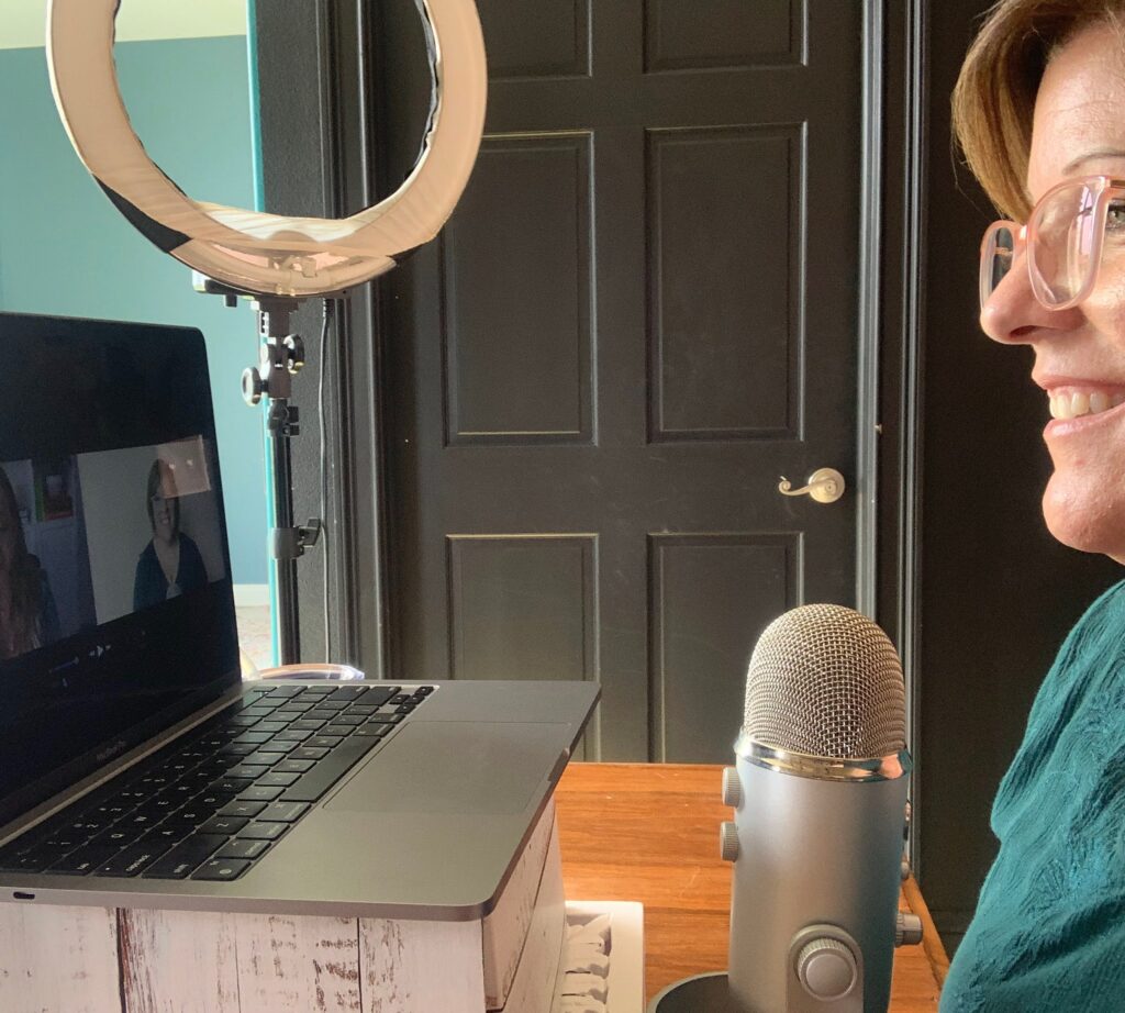 Woman sitting at a desk recording a podcast with a laptop, microphone, and ring light setup.