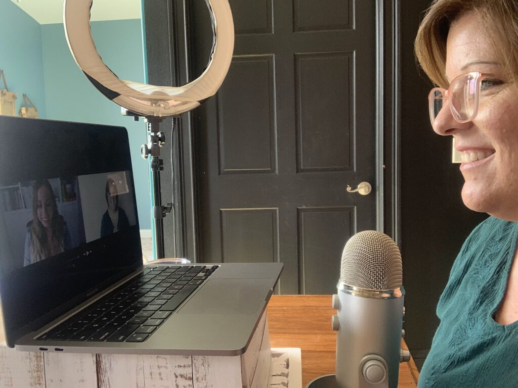 Woman smiling during a virtual meeting, sitting in front of a laptop with a microphone and ring light, showing online business coaching setup.