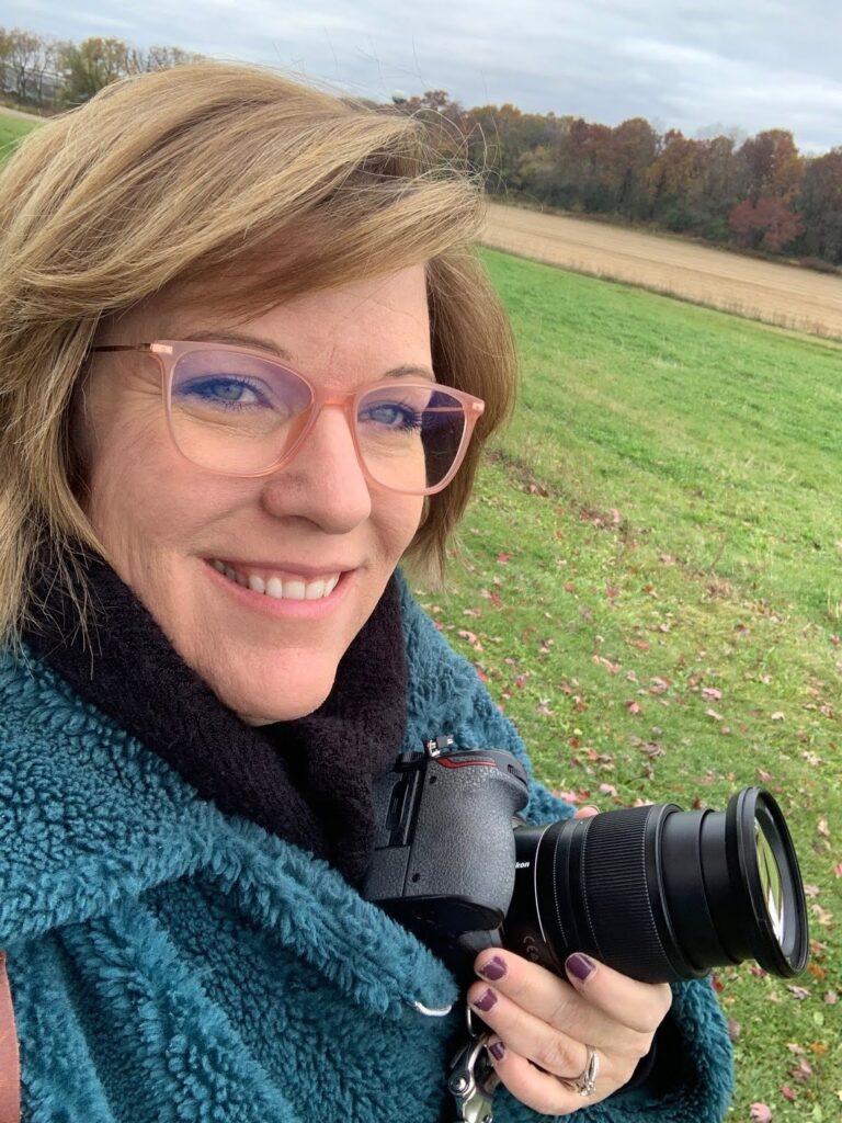 Woman smiling outdoors holding a camera, standing in a grassy field with trees in the background, representing exploring new creative interests.
