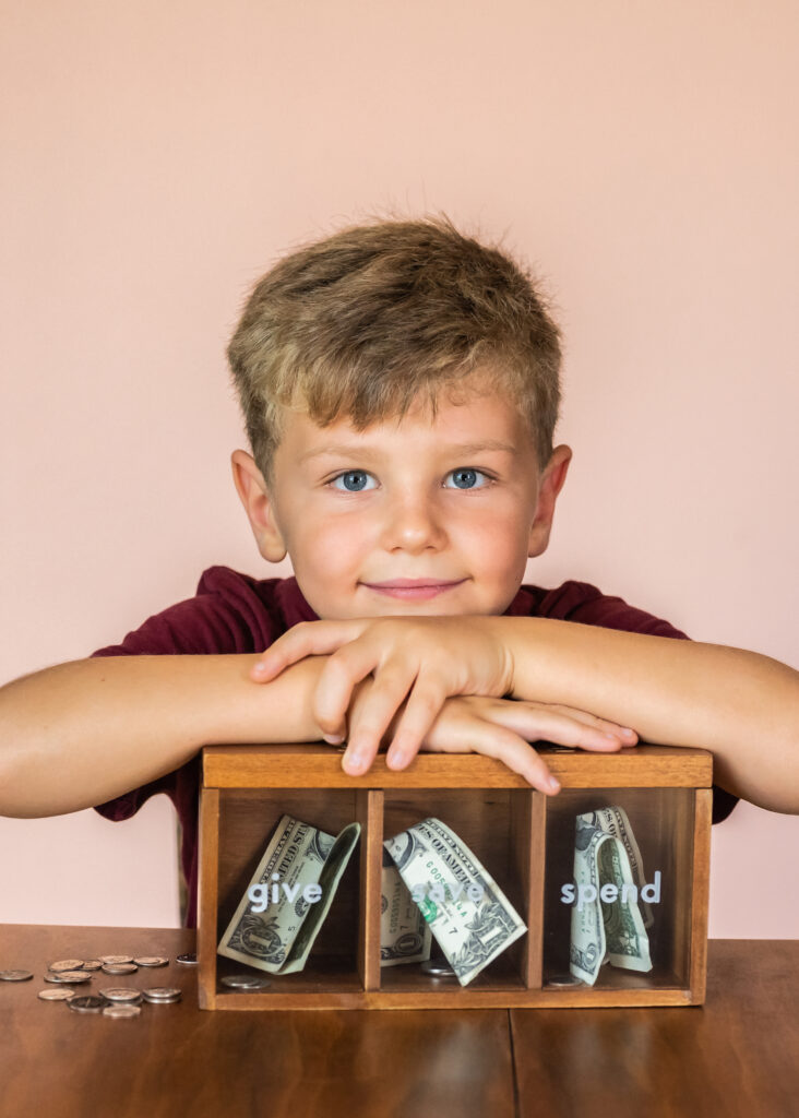 Young boy sitting at a table with a wooden money box labeled give, save, and spend, representing financial education and saving habits.