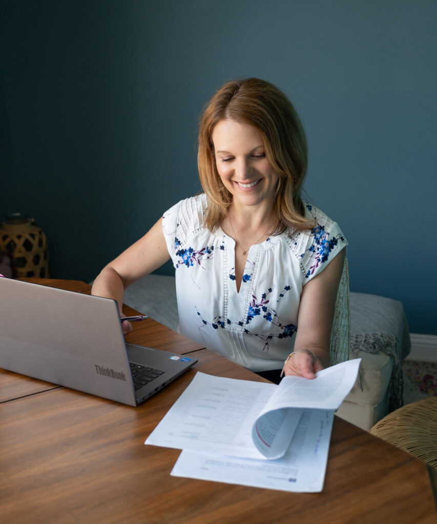 Smiling woman sitting at a wooden table reviewing papers with a laptop in front of her, representing focused business planning and productivity.
