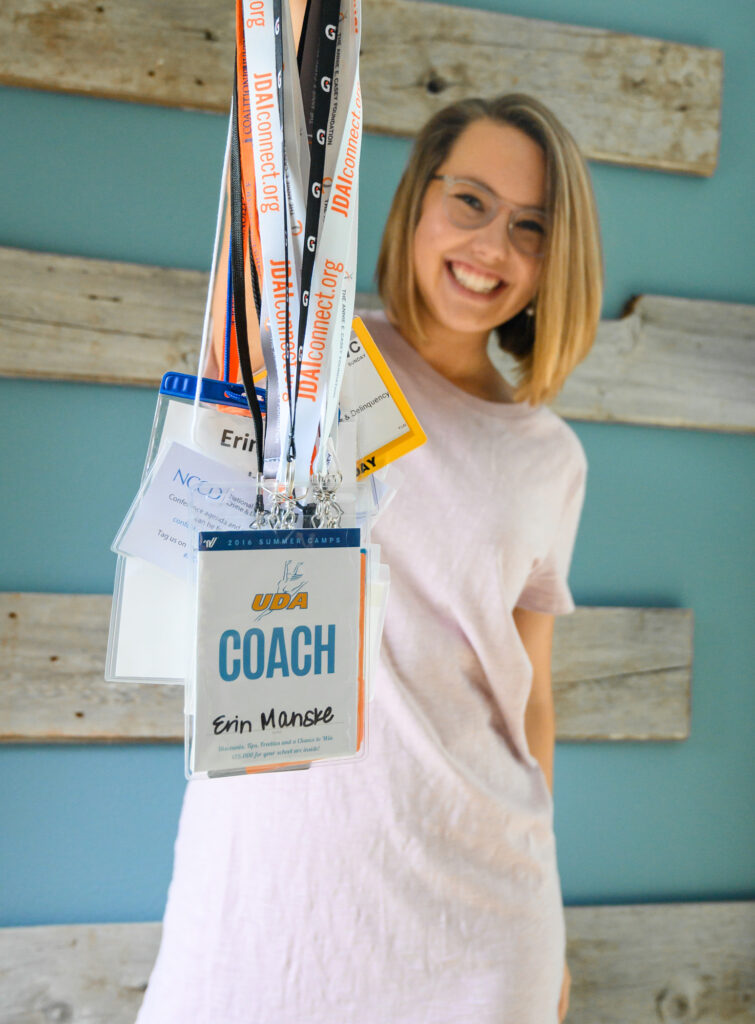 Smiling woman wearing a light pink shirt with multiple coaching lanyards and name badges around her neck, standing against a blue wall with wooden decor