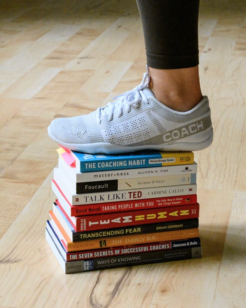Foot wearing a white athletic shoe labeled “Coach” standing on a stack of colorful business, leadership, and coaching books on a wooden floor.