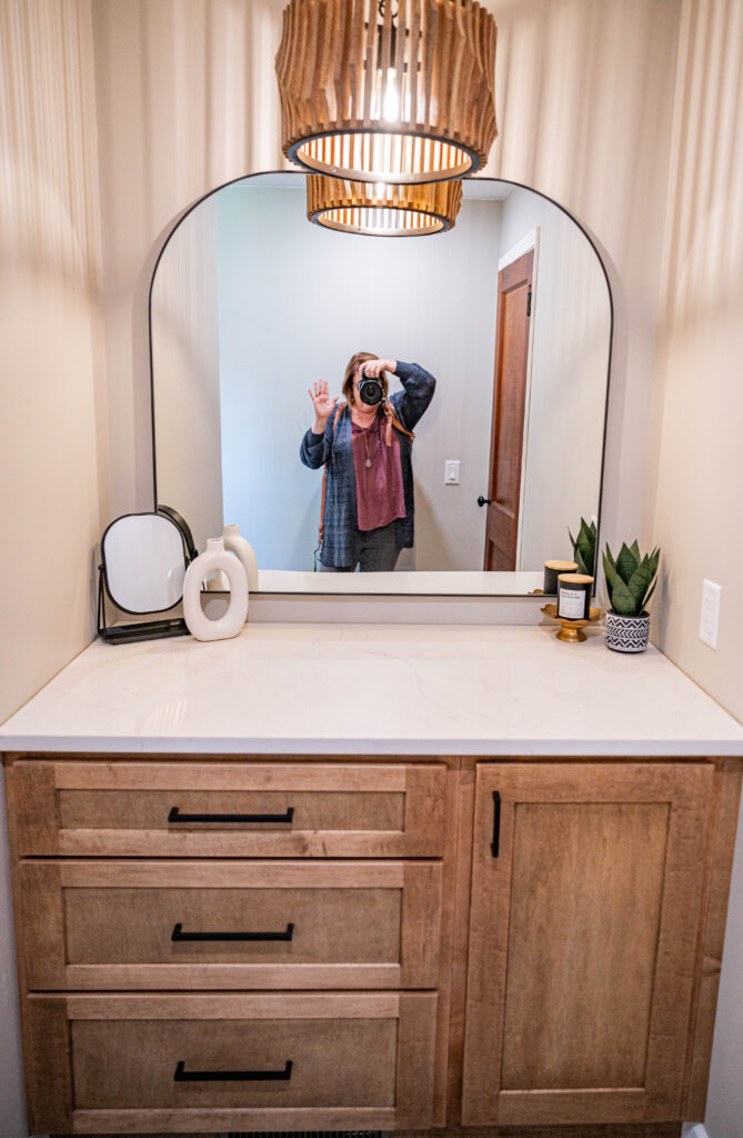 Person taking a photo of themselves in a bathroom mirror, wearing casual clothes with a hanging light above.