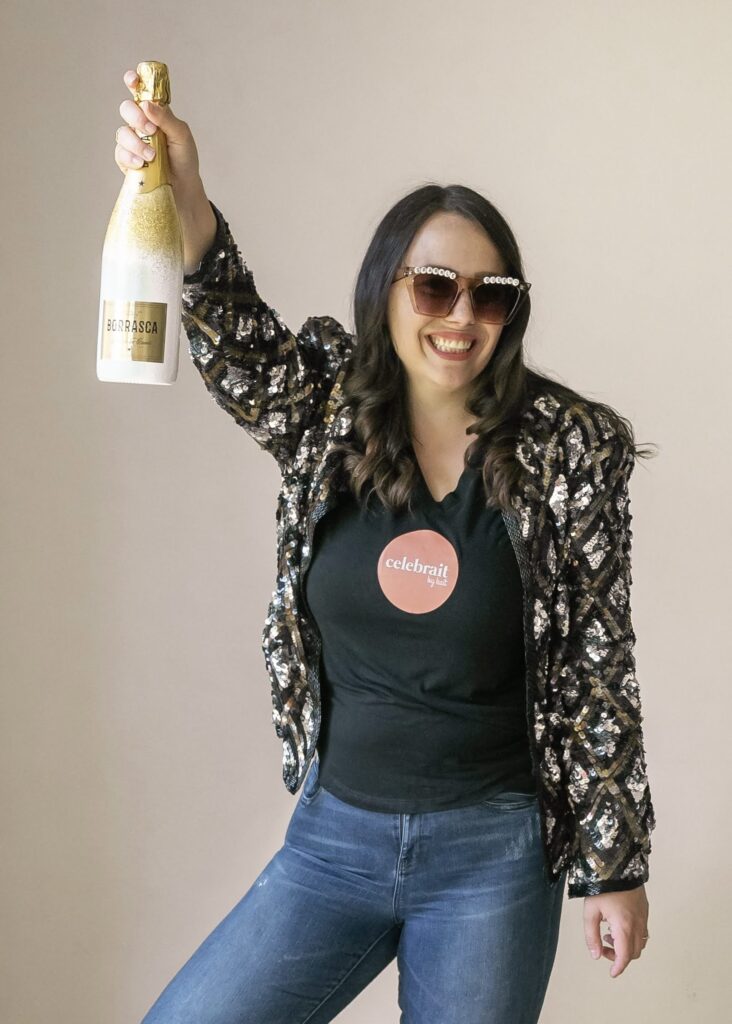 Smiling woman wearing sunglasses, a sequin jacket, and a black t-shirt holding up a bottle of champagne, representing celebration and confidence.