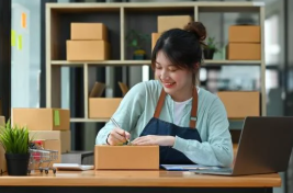 Smiling woman wearing an apron writing on a cardboard box with stacked boxes behind her, representing small business packaging and niche marketing.