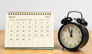 Desk calendar showing March 2022 next to a black analog alarm clock on a wooden surface, representing time management and scheduling.