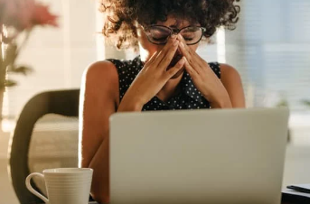Stressed woman with curly hair and glasses sitting at a desk with her laptop, covering her face with her hands due to business overwhelm.