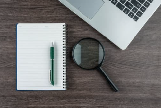 Open notebook with a green pen and black magnifying glass placed on a wooden desk next to a silver laptop, representing research, planning, and analysis tasks.