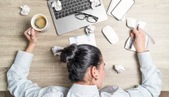 Overhead view of a woman lying her head on a desk near a laptop, coffee cup, and crumpled papers, feeling tired from work.

