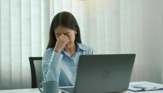 Frustrated woman sitting at a desk with a laptop, holding her forehead with eyes closed, representing marketing overwhelm and stress in business.