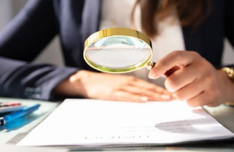 Woman holding a gold magnifying glass over a document on a desk with pens and papers in focus.