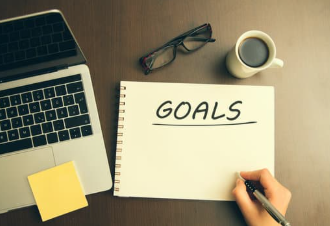 Notebook with the word “GOALS” written on it, placed on a wooden desk beside a laptop, pen, eyeglasses, coffee mug, and sticky note, symbolizing planning and focus.