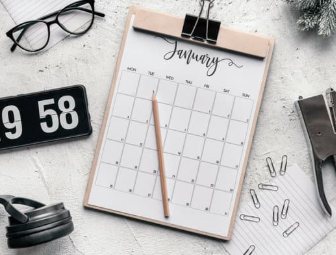Flat lay of January calendar clipboard with pencil, black glasses, clock showing 9:58, and paper clips on white desk.