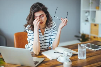 Frustrated woman holding glasses and pinching her nose bridge while working on a laptop at a desk with a glass of water, feeling overwhelmed with marketing tasks.