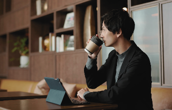 Young man in a black suit sitting in a café, drinking coffee while working on a laptop with shelves in the background, symbolizing stepping back from hustle culture.