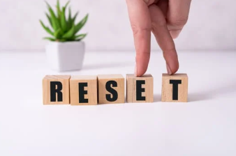 Hand placing wooden block letters spelling “RESET” on a table with a small plant in the background, symbolizing personal and business resets.