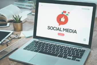 Laptop screen showing social media logo with thumbs up icon, sitting on a wooden desk with a plant and notebook beside it.