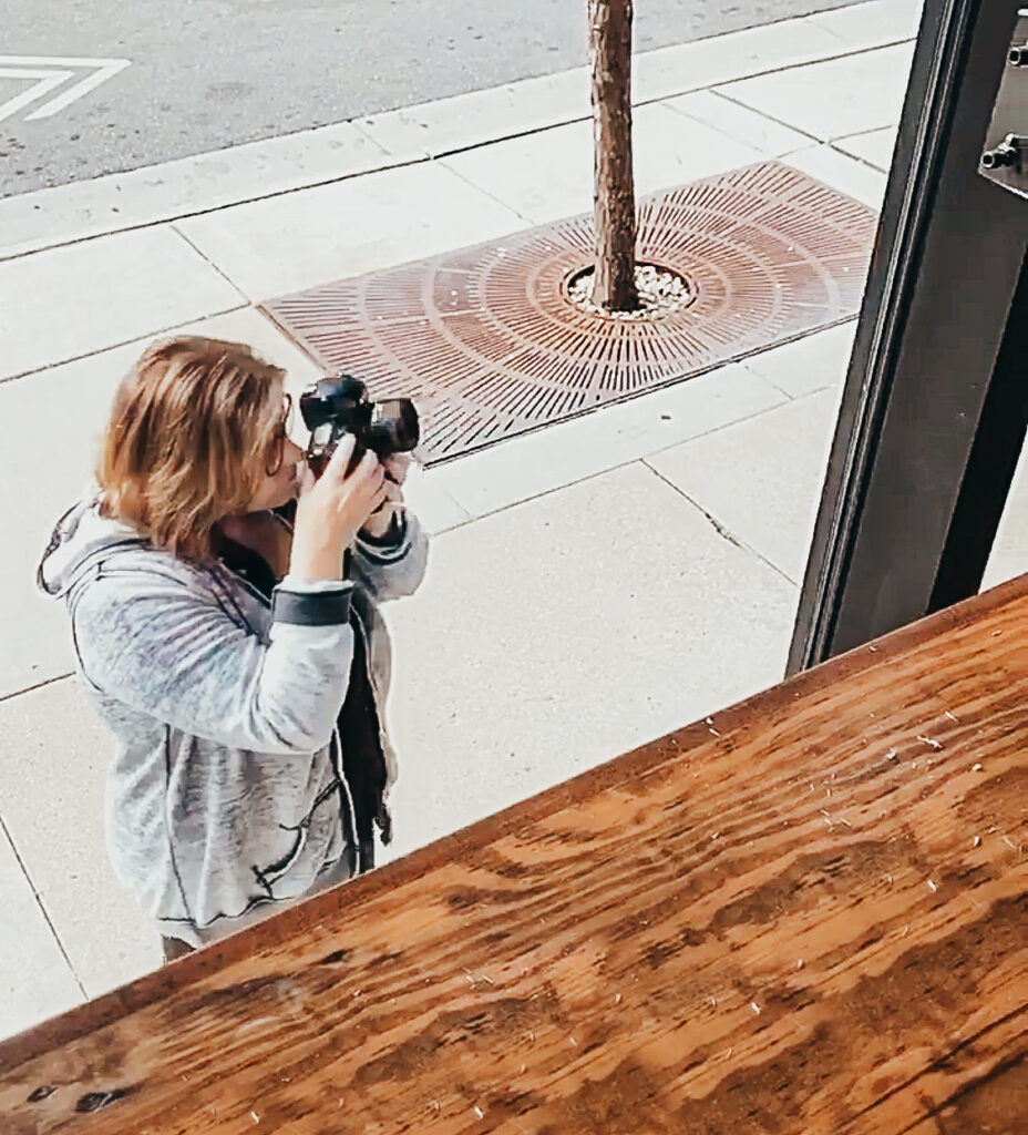 Woman wearing a grey jacket taking photos with a camera from a street sidewalk, viewed from an elevated indoor window.