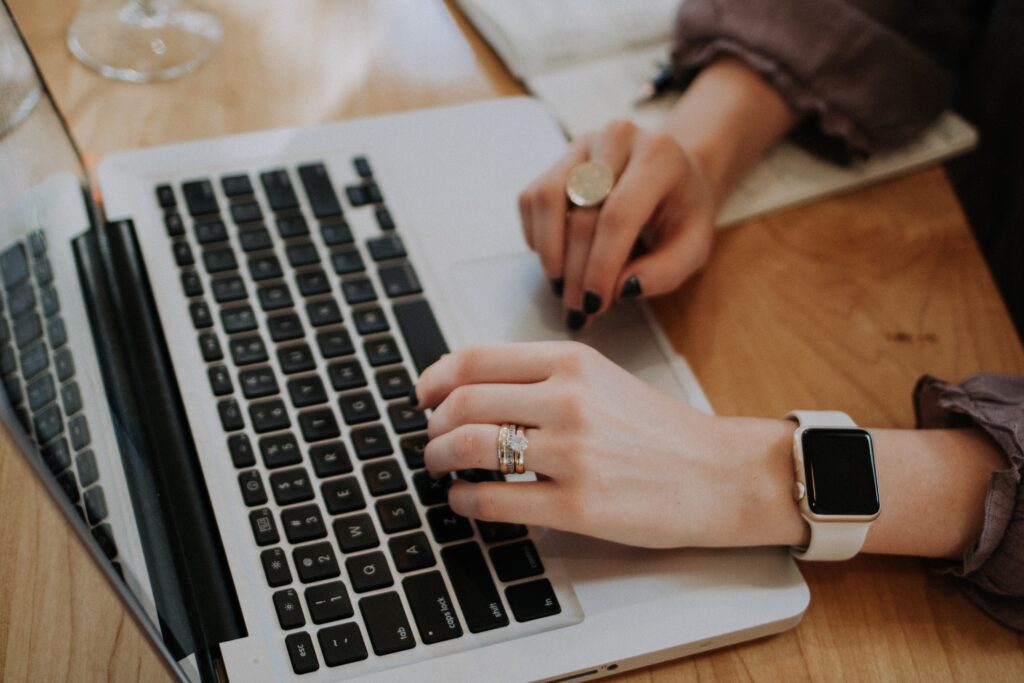 Woman typing on laptop with smartwatch and rings visible on hand.