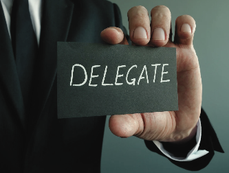 Man in a black suit holding a black card with the word “DELEGATE” written on it, symbolizing delegation in solopreneurship.