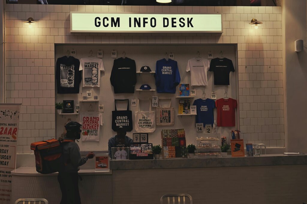 Person wearing a helmet and backpack standing at GCM info desk with merchandise display, including shirts, hats, and souvenirs on a tiled wall.