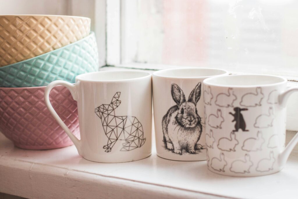 Three white mugs with different rabbit designs placed on a windowsill next to stacked textured bowls in pastel colors, representing cozy kitchenware and home decor.