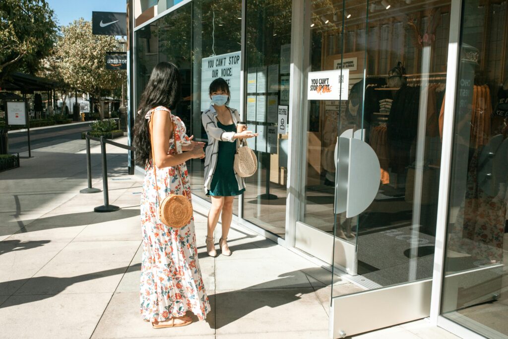 Two women standing outside a store entrance, one wearing a floral dress and the other wearing a mask and green dress, representing shopping and pandemic safety.