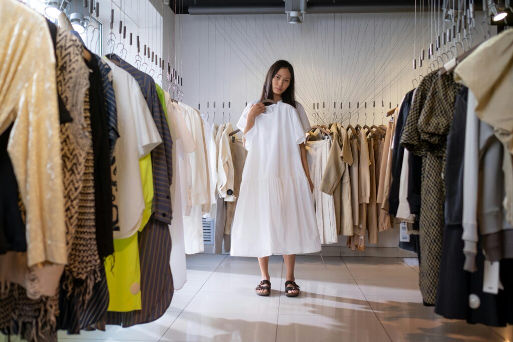 Woman standing in a clothing store holding a white dress surrounded by racks of clothes, representing shopping and fashion choices.