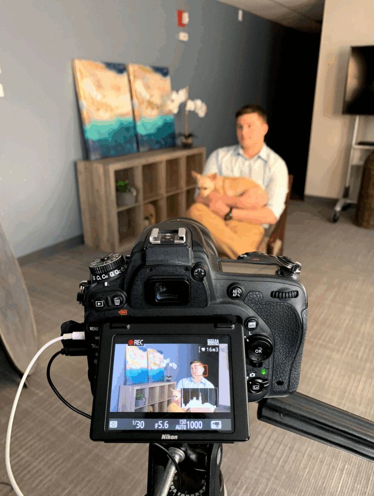 Behind-the-scenes view of a video shoot capturing a man holding a small dog during an interview, with the camera display showing a focused recording of the subject seated in a well-lit office space.