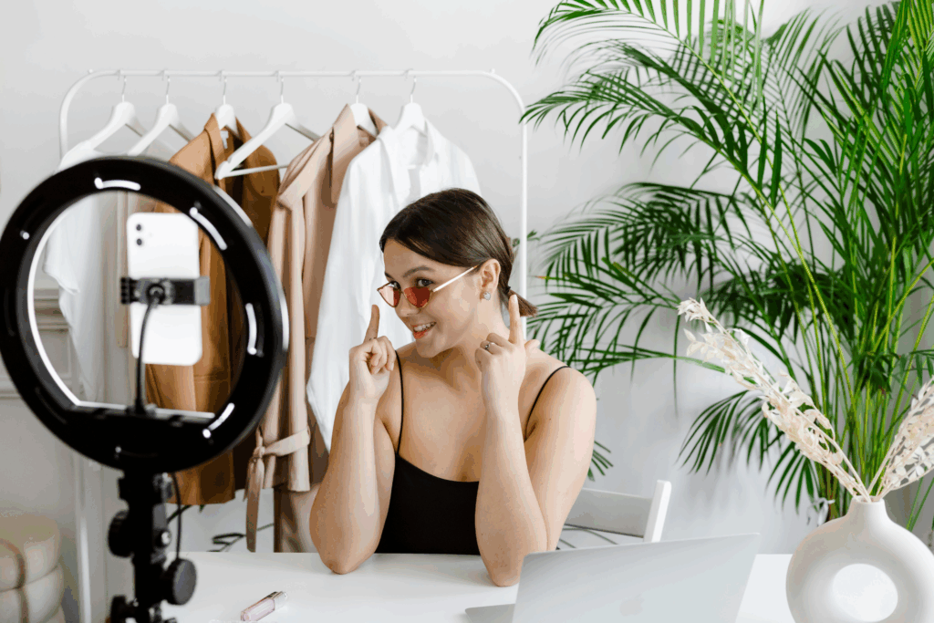 Young woman recording a fashion and lifestyle video using a smartphone and ring light, showcasing pink sunglasses in a bright, modern home studio with clothing rack and greenery in the background.
