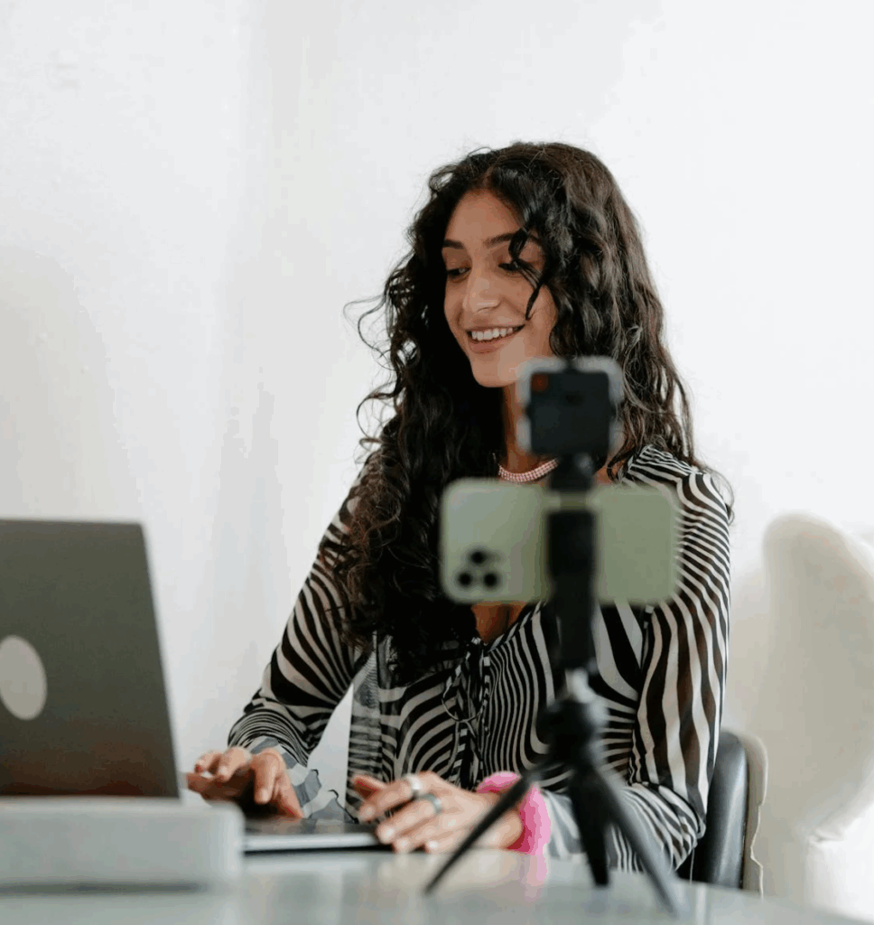 Smiling content creator livestreaming or recording a video while working on a laptop, with a smartphone mounted on a tripod in front.