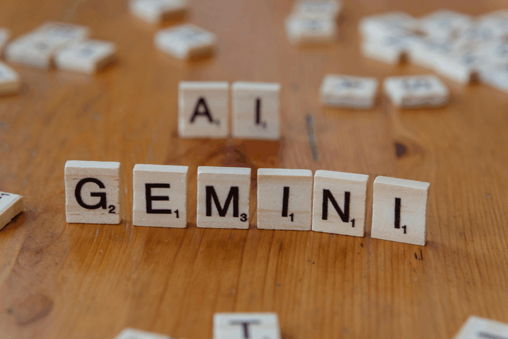 Close-up of Scrabble tiles spelling out "GEMINI" on a wooden surface, with "AI" tiles blurred in the background, suggesting a connection between Gemini and artificial intelligence.